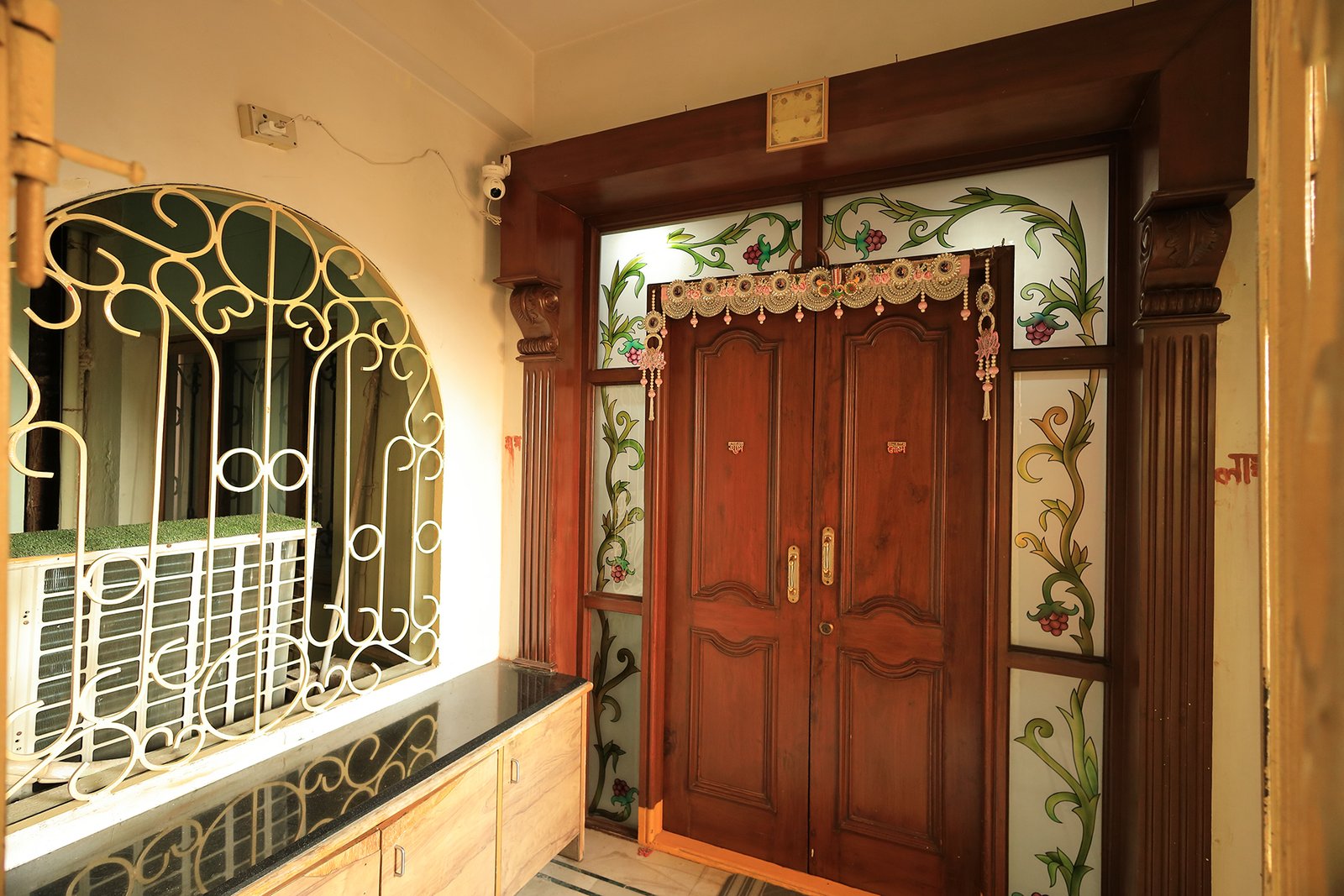 Main wooden door with hand-painted glass panels and a traditional torana, with the wooden shoe cabin beside it